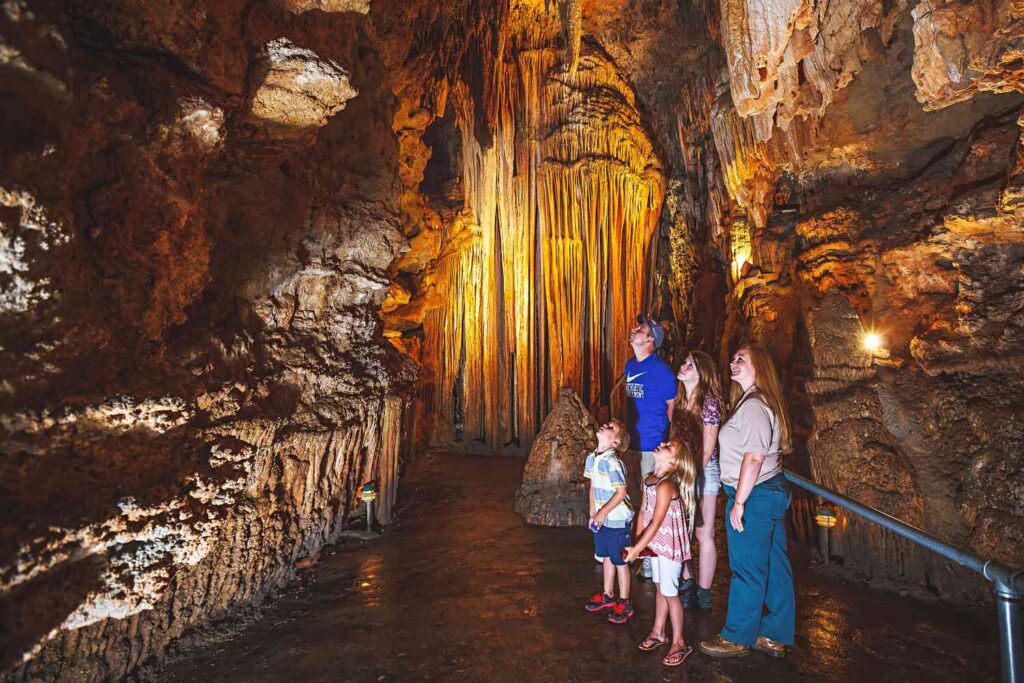 A family explores a dramatic cave filled with towering stalactites and illuminated rock formations in the Ozarks region of Missouri. The golden glow from inside the cavern highlights the natural beauty and geological wonder found near the Lake of the Ozarks. This underground experience showcases a different side of the Missouri Ozarks, where families can enjoy both lake activities and unique attractions in the heart of the Ozarks in Missouri. 