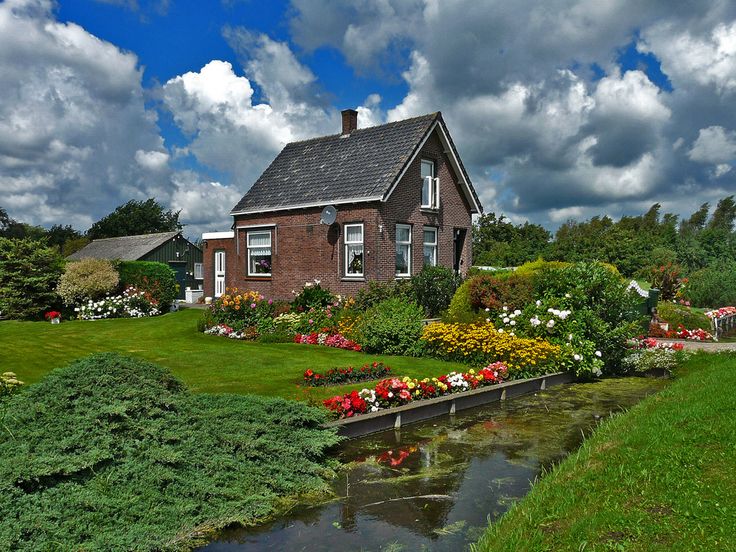 A charming brick house surrounded by a lush, vibrant garden with colorful flowers, a small canal running in front, and a dramatic, partly cloudy sky overhead.