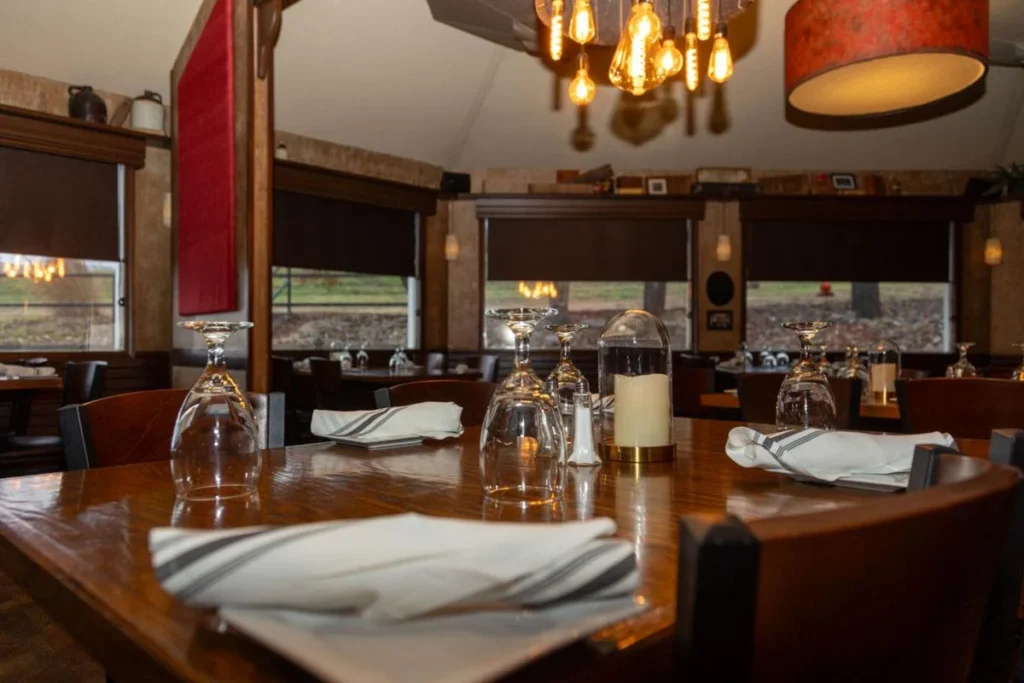  A rustic yet elegant restaurant near Lake of the Ozarks, Missouri, with wooden tables set for dinner, upside-down wine glasses, folded napkins, and warm pendant lighting. Large windows offer a view of the surrounding Ozarks landscape, creating an inviting atmosphere for guests enjoying a meal in the heart of the Missouri Ozarks. A great spot to relax after exploring the Ozark lakes or spending the day on the water at Lake of the Ozarks MO.