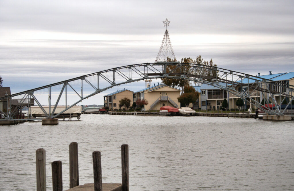  A small arched footbridge spans a calm waterfront canal lined with lake houses and boat docks near Lake Erie, Ohio. A large metal Christmas tree structure rises behind the houses, adding a festive touch to this lakeside town. Popular among anglers and vacationers alike, this scene reflects life in Ohio lake communities known for lakefront living, scenic views, and access to Lake Erie’s best fishing spots, especially for those following Ohio lake fishing events and tournaments.
