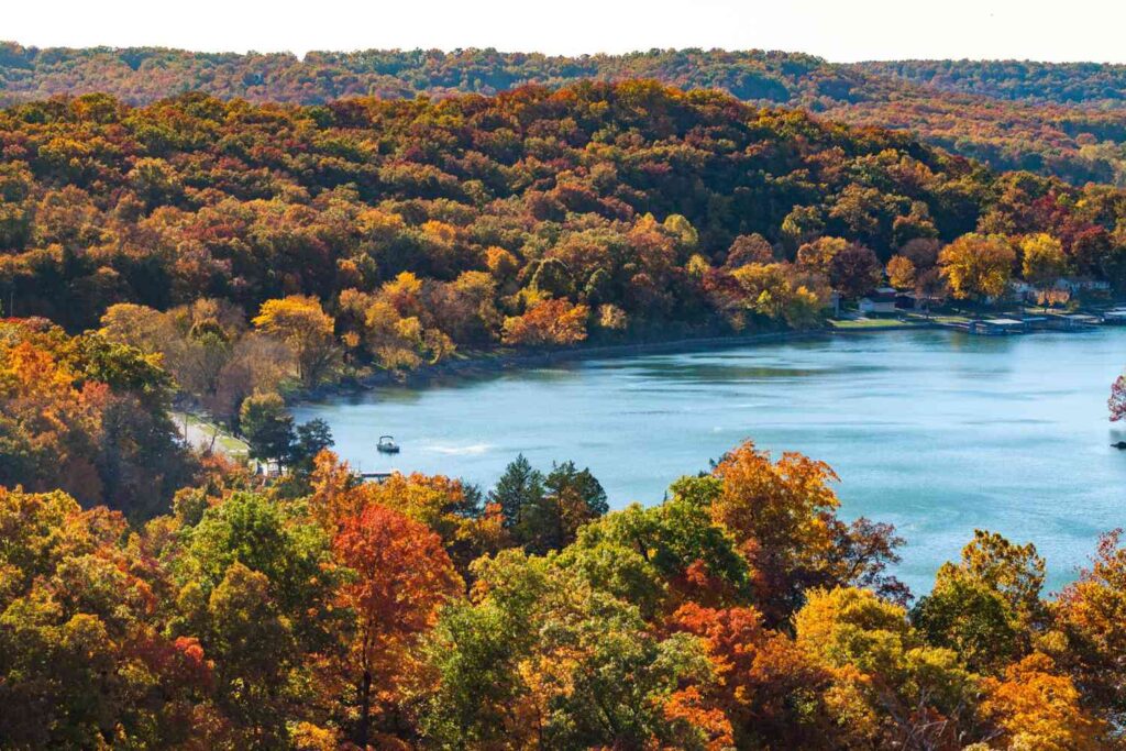 A peaceful lake surrounded by a forest bursting with vibrant autumn colors, including deep reds, oranges, yellows, and greens. A small boat glides across the calm water, while rolling hills covered in colorful trees stretch into the distance under a soft, hazy sky. The scene captures the beauty of fall in a quiet, natural setting. 