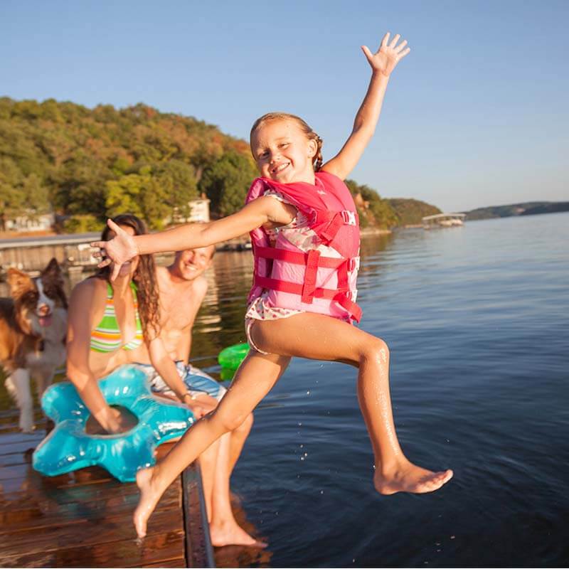 A smiling young girl in a pink life jacket joyfully leaps off a dock into the water at Lake of the Ozarks in Missouri, with her arms raised mid-air. Behind her, family members relax on pool floats and enjoy the sunshine, with a forested shoreline and lakeside homes in the background. This lively summer scene highlights the fun and family-friendly atmosphere of the Lake of the Ozarks MO, one of the most popular vacation spots in the Missouri Ozarks. 