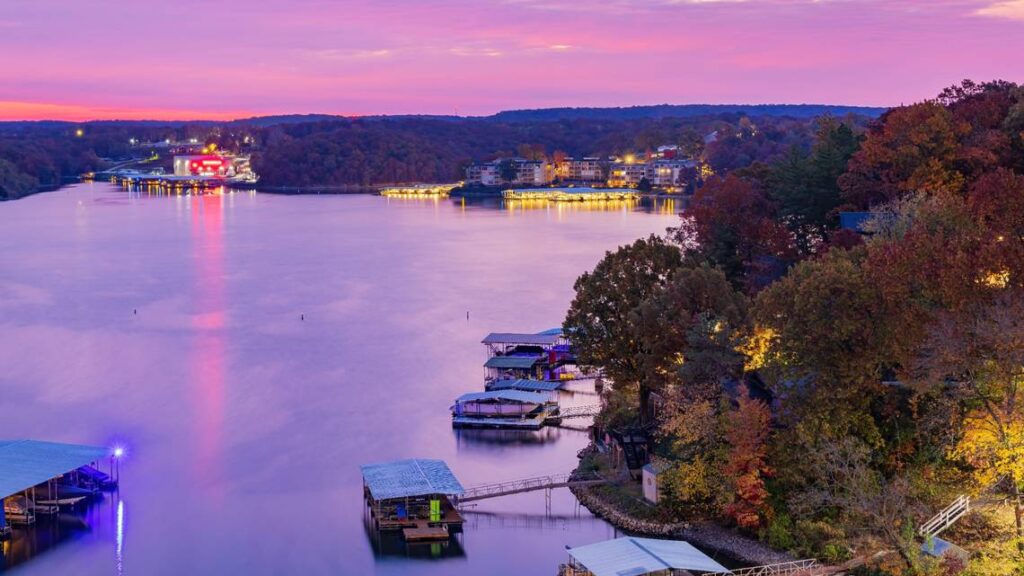  A peaceful evening at Lake of the Ozarks in Missouri, with docks and boathouses glowing under soft purple and pink sunset skies. Lights from nearby restaurants and lakeside homes reflect off the calm water, while autumn foliage lines the hills of the Missouri Ozarks. This tranquil lakeside view showcases the beauty of the Ozarks in Missouri and makes Lake Ozarks MO a popular destination for sunset cruises, getaways, and lakeside dining.