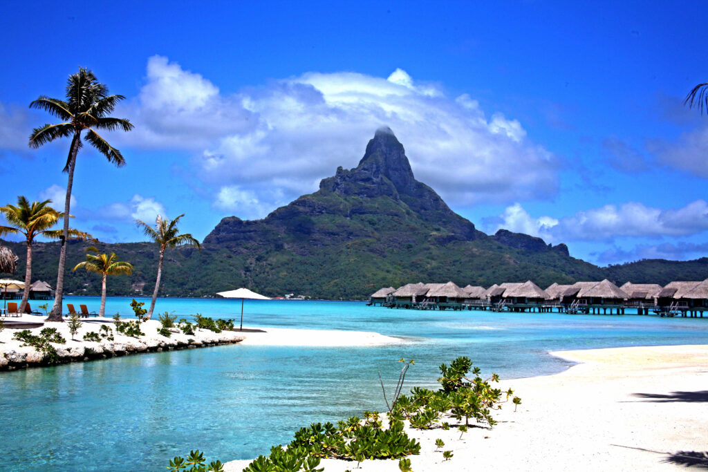 Overwater bungalows on turquoise waters beneath the dramatic peak of Mount Otemanu in Bora Bora, French Polynesia, surrounded by palm trees and tropical scenery.