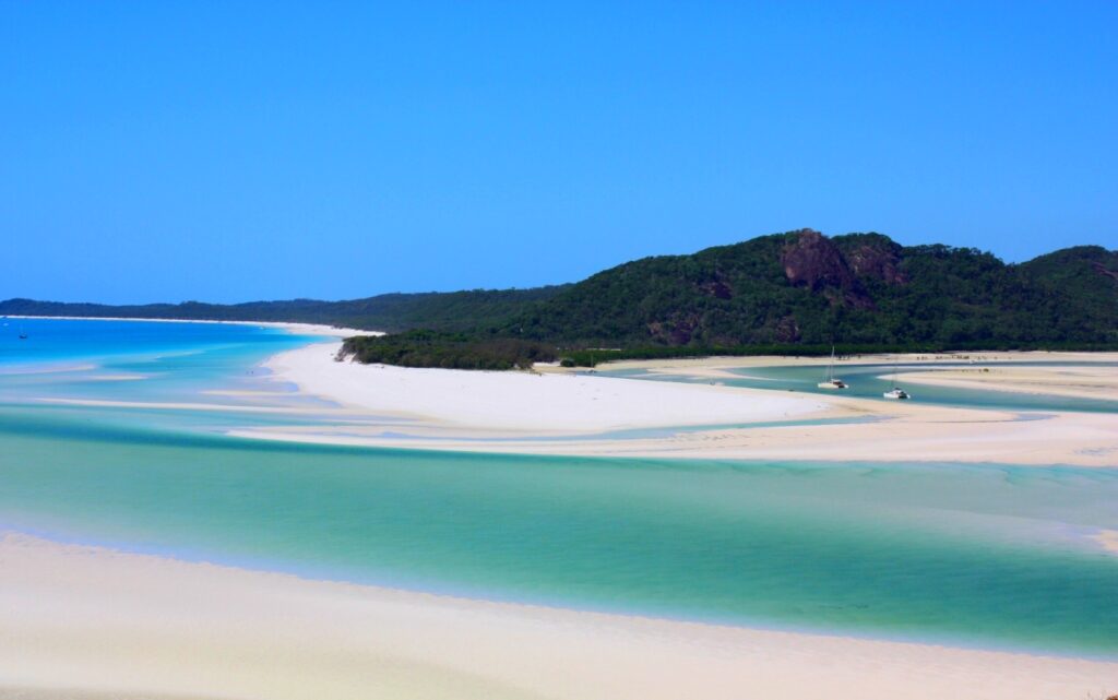 Whitehaven Beach in the Whitsunday Islands, Australia, featuring swirling white silica sand bars and vibrant turquoise waters with yachts anchored in the shallows, bordered by lush forested hills.