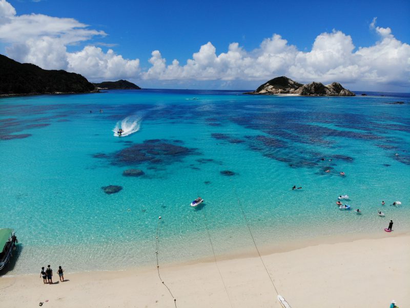 Aharen Beach on Tokashiki Island in Okinawa, Japan, featuring clear turquoise waters, coral reefs, and beachgoers enjoying swimming and snorkeling under a sunny blue sky.