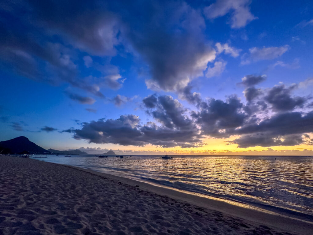 Sunset at Flic en Flac Beach in Mauritius with golden hues on the horizon, dark clouds scattered across the sky, calm rippling waves, and a peaceful sandy shoreline under twilight.