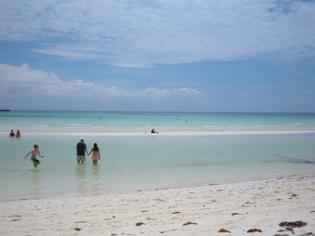 Visitors wade into the shallow, crystal-clear waters of Gold Rock Beach in Grand Bahama, Bahamas, surrounded by soft white sand and a tranquil turquoise sea under a partly cloudy sky.