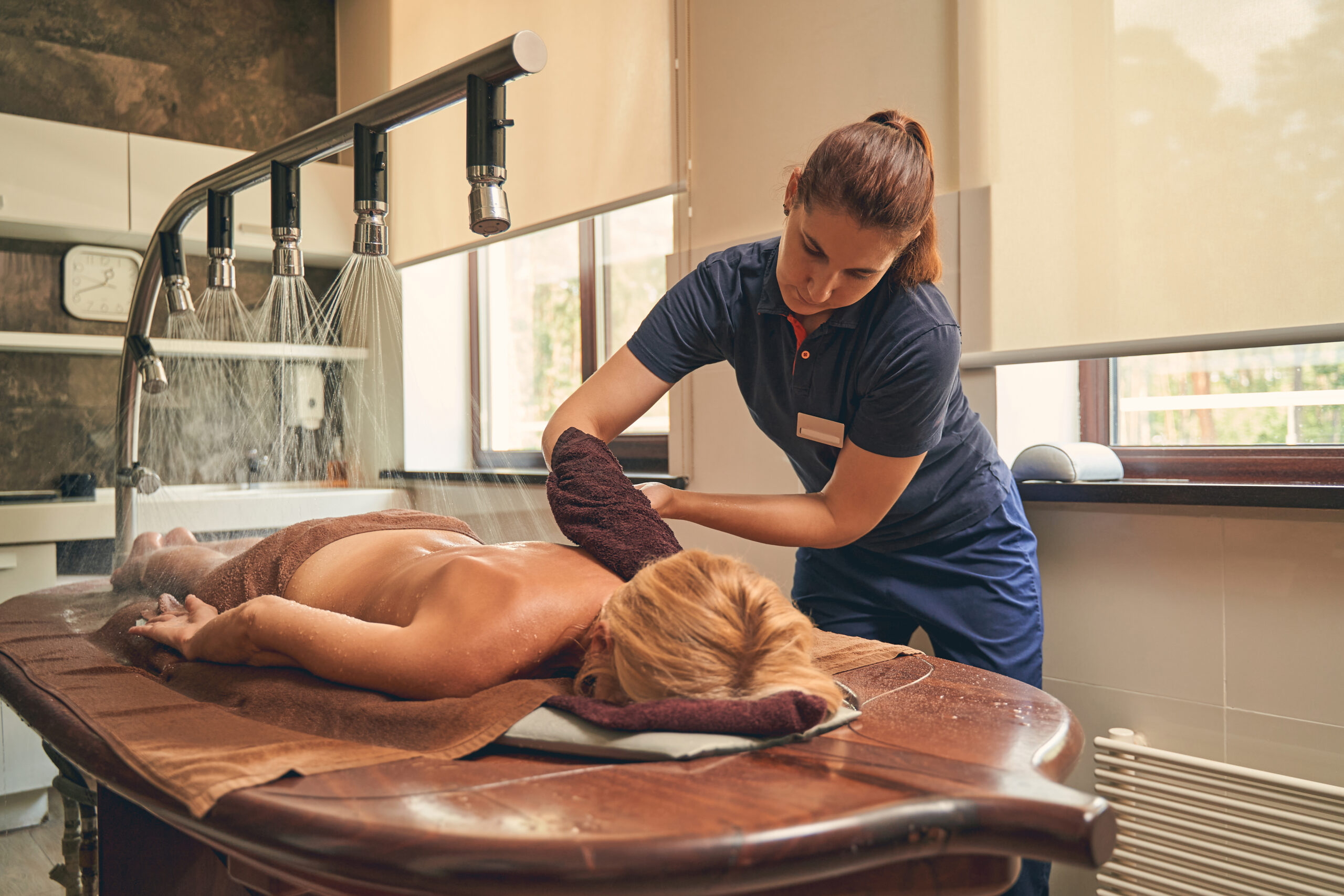 Spa therapist performing a thalassotherapy water massage on a woman lying on a wet treatment table in a wellness center in Greece