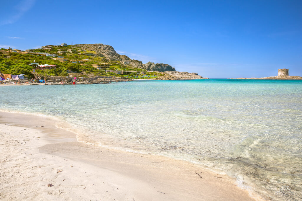 La Pelosa Beach in Stintino, Sardinia, featuring transparent shallow waters, soft white sand, rocky hillsides, and a distant Aragonese tower under a clear blue sky.
