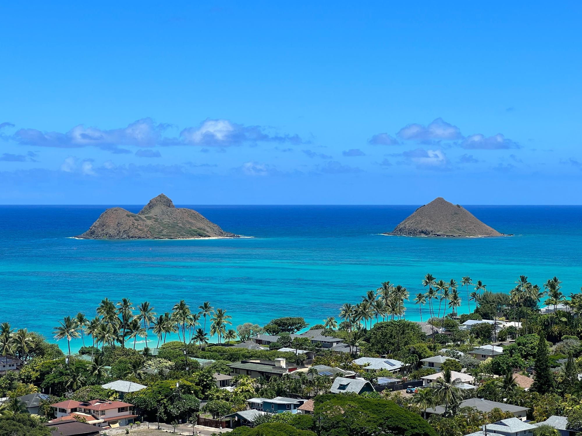 Aerial view of Lanikai Beach in Oahu, Hawaii, with turquoise waters, white sand, lush palm trees, and the twin Mokulua Islands in the distance under a clear blue sky.