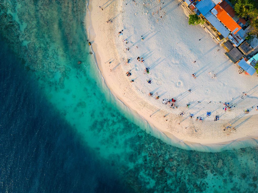 Aerial view of White Beach in Moalboal, Cebu, Philippines, showing a curved shoreline with soft white sand, turquoise waters with visible coral formations, and people relaxing near beach huts and vibrant rooftops.