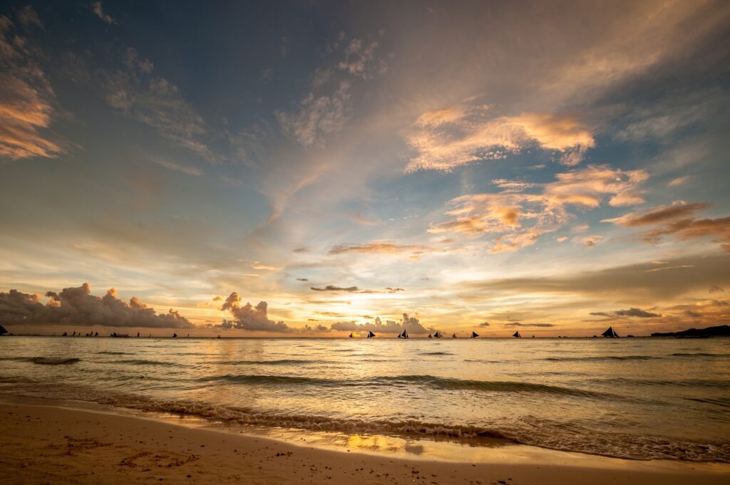 Golden sunset over the calm waters of Boracay Beach in the Philippines, with soft clouds scattered across a colorful sky and traditional paraw sailboats silhouetted on the horizon.