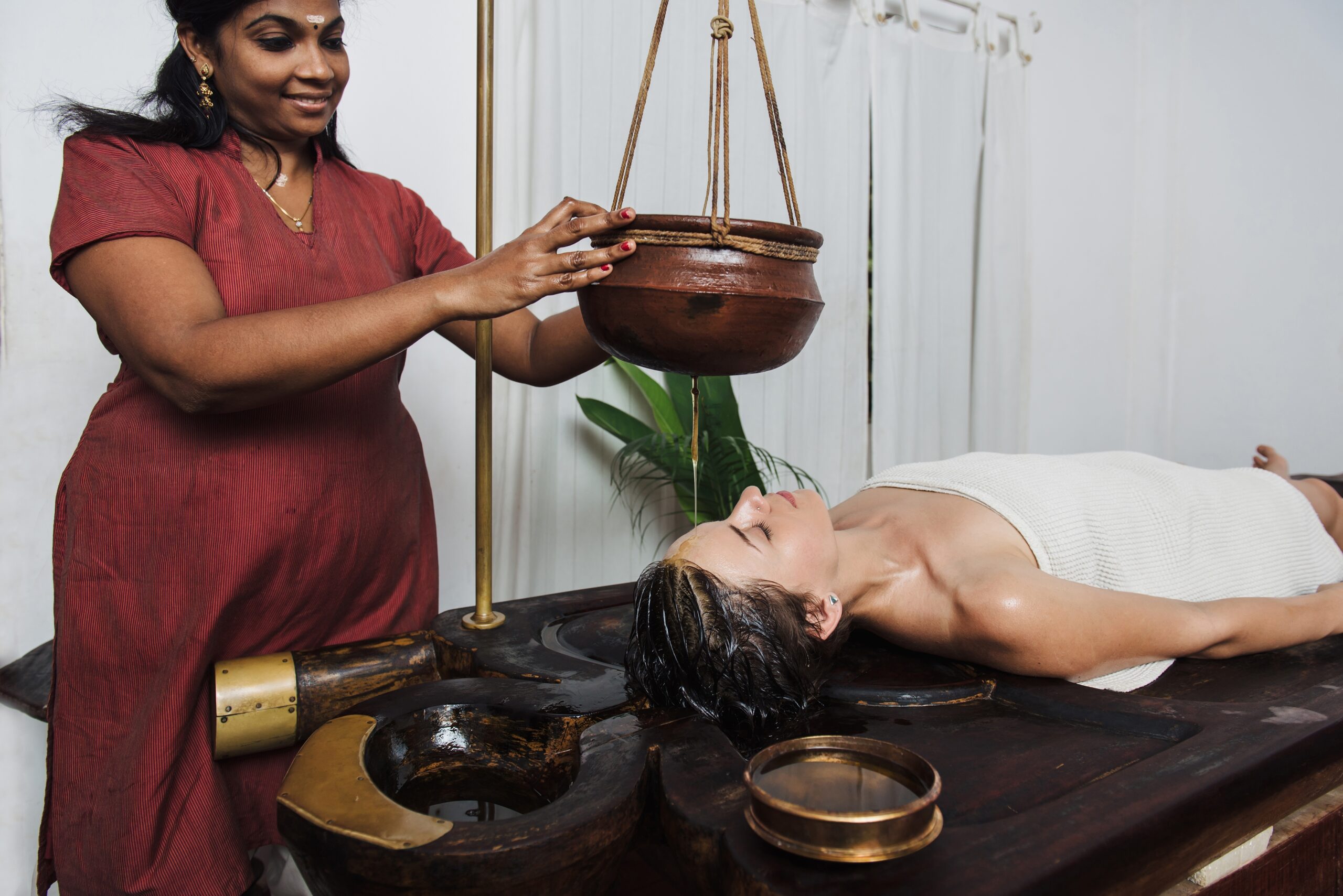 Ayurvedic practitioner performing Shirodhara therapy on a woman lying on a traditional wooden table as warm oil streams onto her forehead