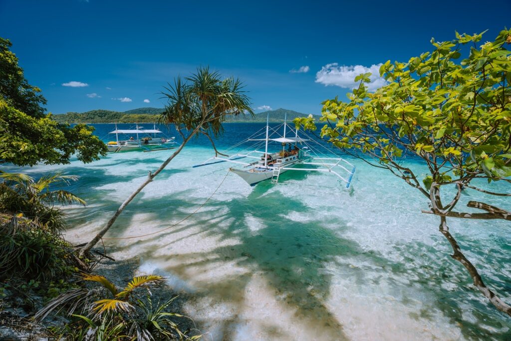 A traditional Filipino outrigger boat docked in the shallow turquoise waters of El Nido, Palawan, surrounded by tropical foliage, white sand, and distant forested islands under a vibrant blue sky.