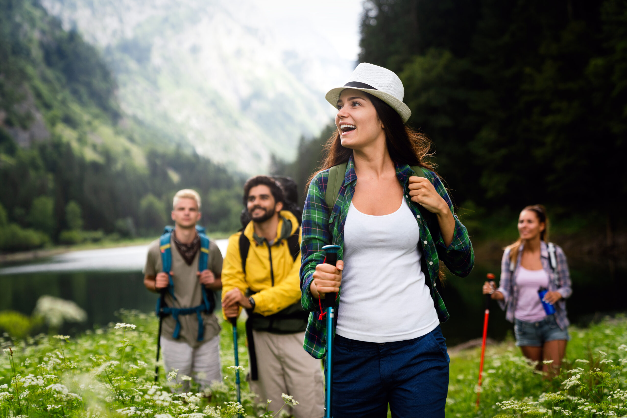 Happy group of young adults hiking with backpacks and trekking poles through a lush green mountain valley