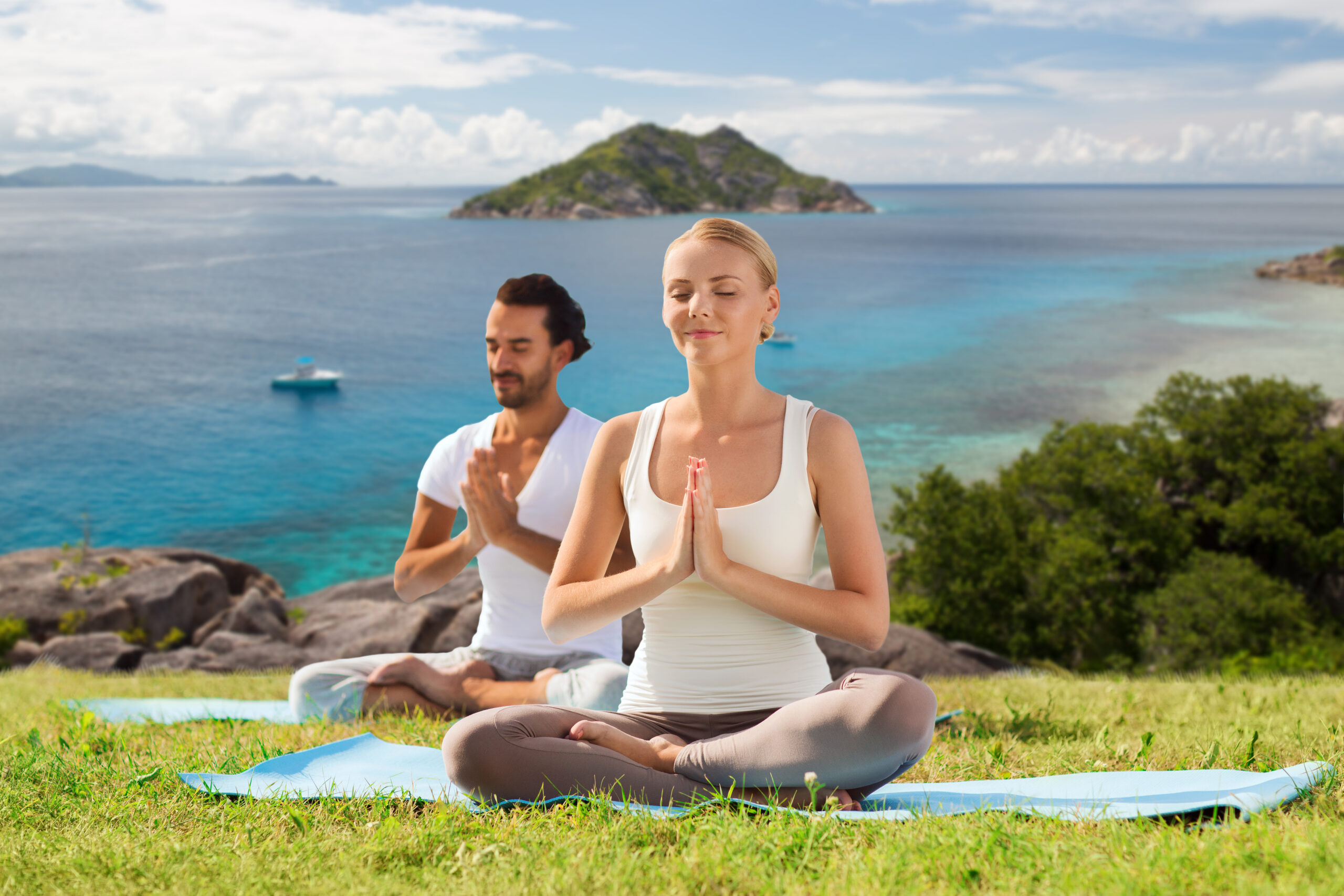 Man and woman meditating in a seated yoga pose on grass with ocean and island in the background