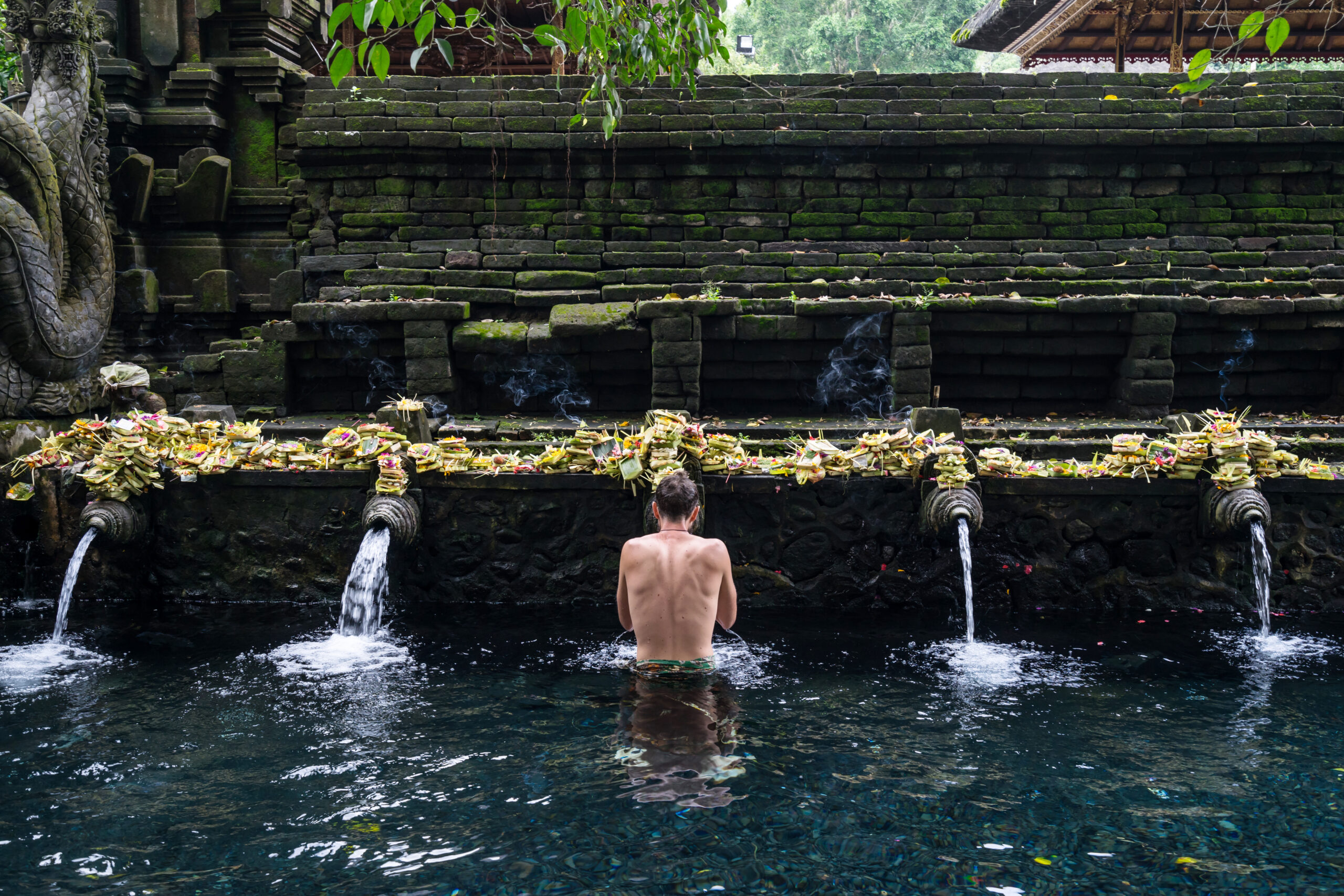Man performing purification ritual at Tirta Empul holy spring temple in Bali, surrounded by stone fountains, incense smoke, and traditional offerings.