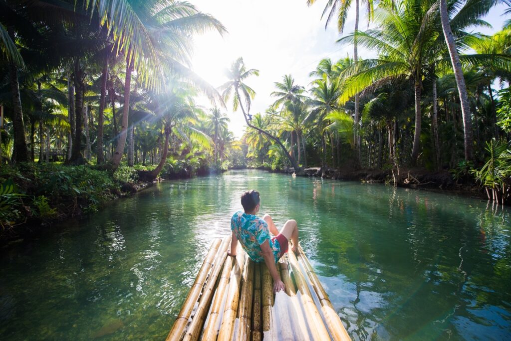 A traveler relaxes on a bamboo raft drifting along the Maasin River in Siargao, Philippines, surrounded by towering coconut palms, lush tropical greenery, and calm green waters under soft morning sunlight.
