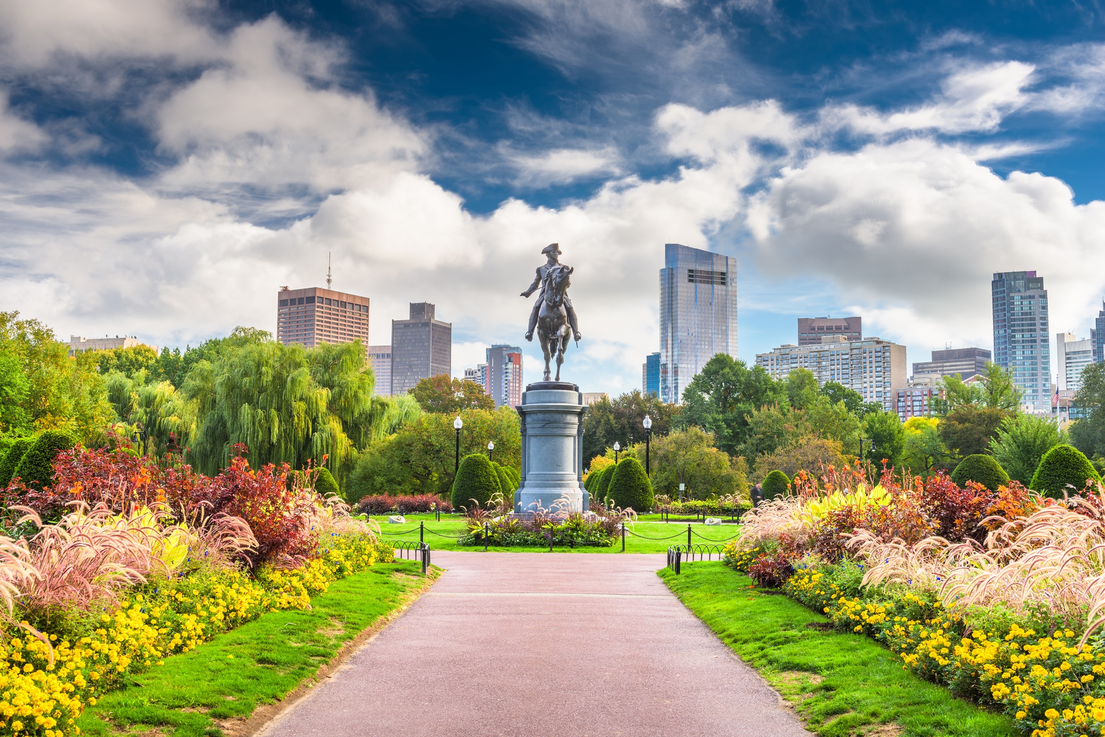 Statue of George Washington in Boston Public Garden surrounded by vibrant spring flowers, with the city skyline in the background under a partly cloudy sky.