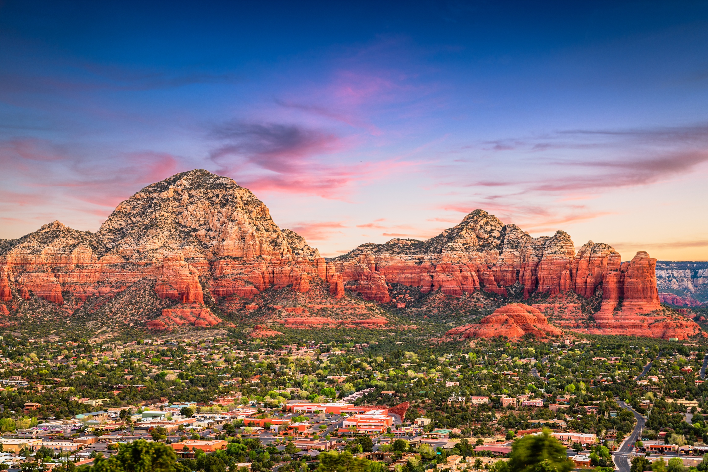 Panoramic view of Sedona, Arizona at sunset with red rock formations, desert vegetation, and the town below under a colorful sky of pink and blue hues.