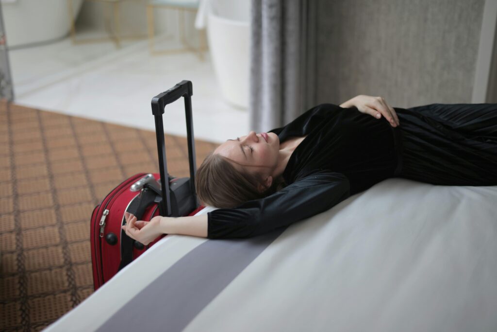 Woman in a black travel outfit lying exhausted on a hotel bed beside a red suitcase — capturing jet lag fatigue and the need for post-flight rest.