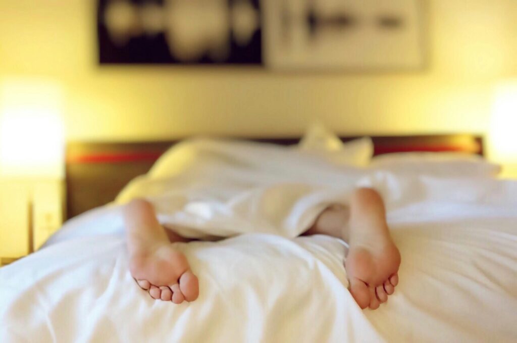 Blurred image of a person’s bare feet peeking out from under white hotel bedding, symbolizing restful sleep, hotel stays, and spontaneous travel comfort.