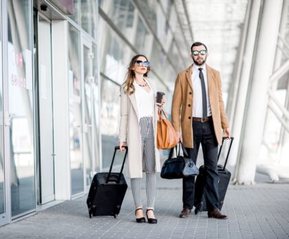 A stylish couple in sunglasses and coats walking outside an airport with luggage, representing luxury travel on a budget with Travel Crock.