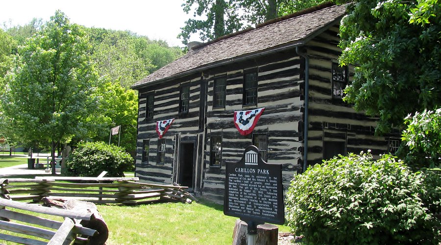 Two-story black-and-white log cabin with patriotic bunting at Carillon Historical Park surrounded by greenery and a wooden fence