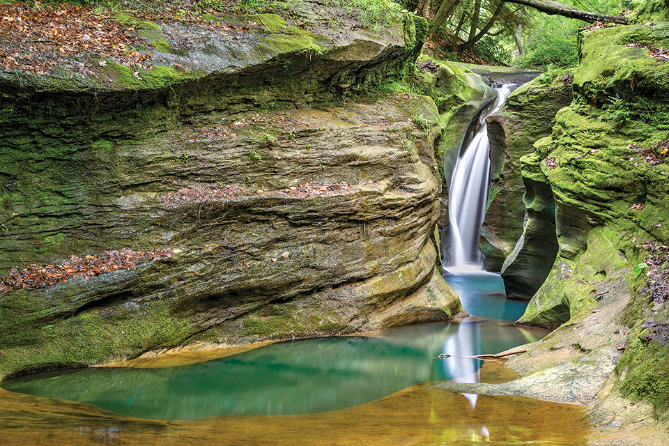 Narrow waterfall cascading into a turquoise pool between moss-covered rock walls in a lush forest setting