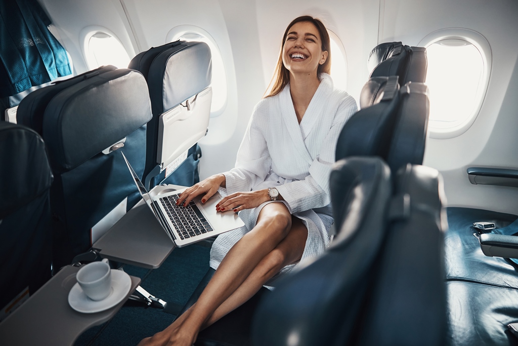 Happy woman in a bathrobe using a laptop and enjoying coffee in a business class airplane seat—illustrating affordable premium flying with Travel Crock.