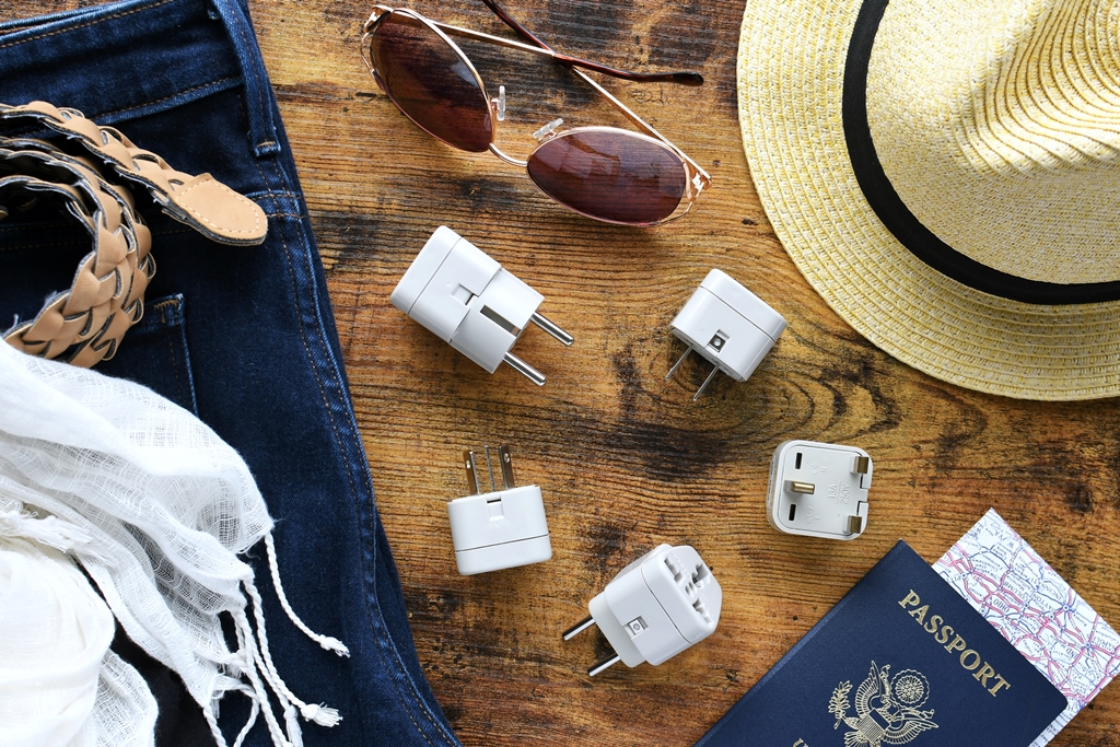 Flat lay of travel essentials including universal power adapters, sunglasses, hat, passport, jeans, and scarf—perfect for Travel Crock trips