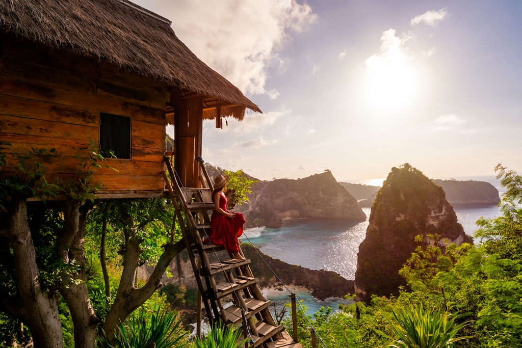 Woman in red dress sitting on stairs of a treehouse overlooking a tropical cliffside ocean view—showcasing budget luxury travel with Travel Crock.