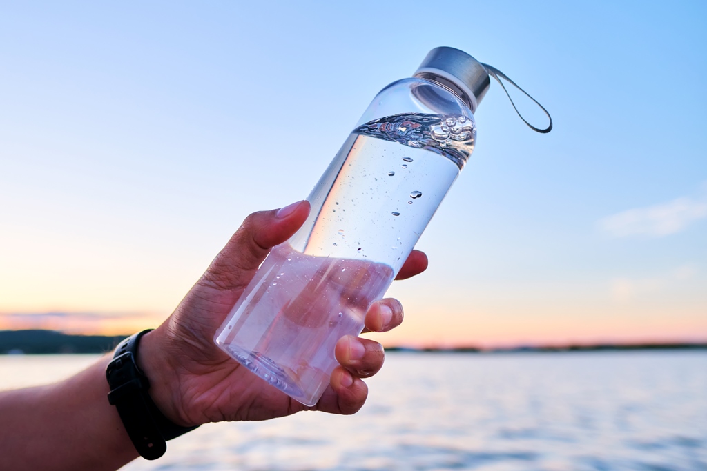Traveler holding a clear refillable water bottle against a sunset backdrop—hydration essential for long flights by Travel Crock.