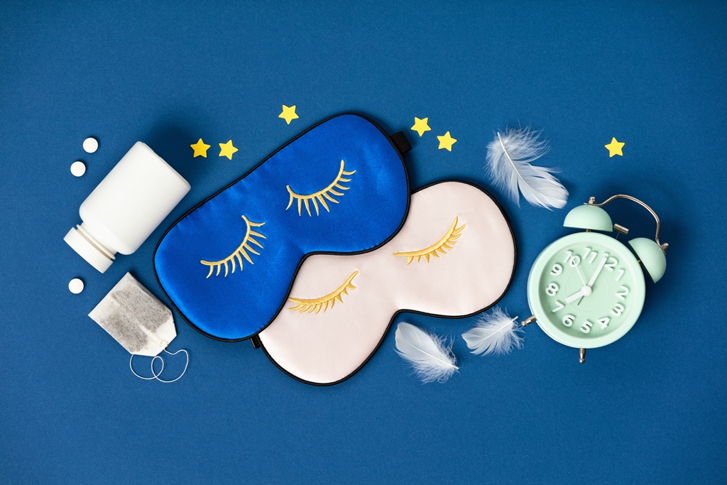 Flat lay of blue and pink eye masks, sleeping pills, herbal tea, feathers, and an alarm clock—ideal sleep kit for long flights by Travel Crock.