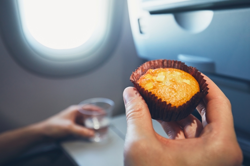 Close-up of a passenger holding a muffin and a cup of water on an airplane tray table—smart snack choice for long flights by Travel Crock.