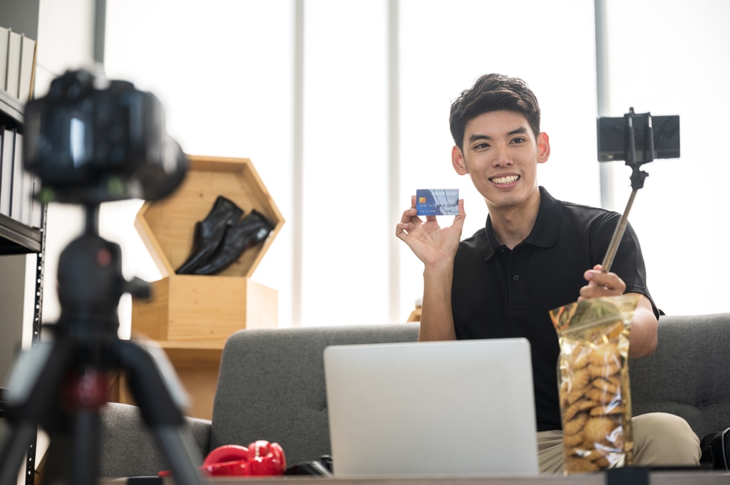 "Smiling young man filming an affiliate product review with a smartphone and camera, holding a credit card and snack bag while working remotely from home"