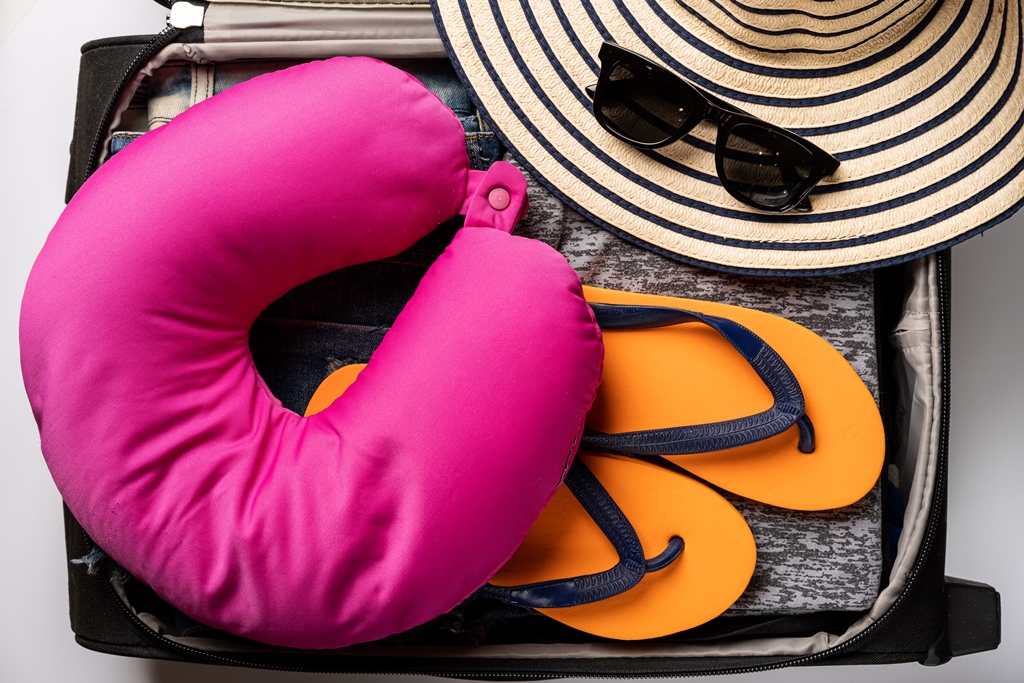 Open suitcase packed with a pink neck pillow, orange flip-flops, striped sunhat, and sunglasses—long flight essentials by Travel Crock.